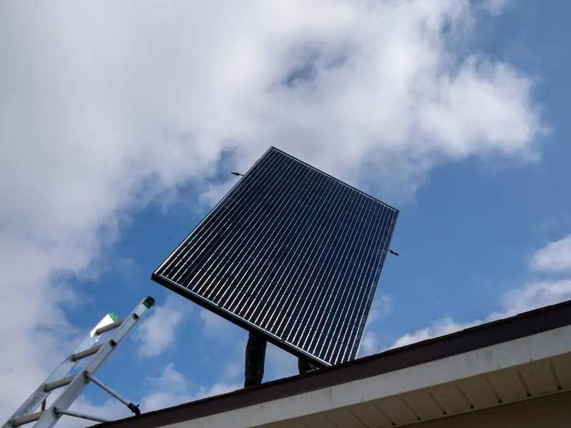 A solar panel being lifted onto a roof against a blue sky with clouds